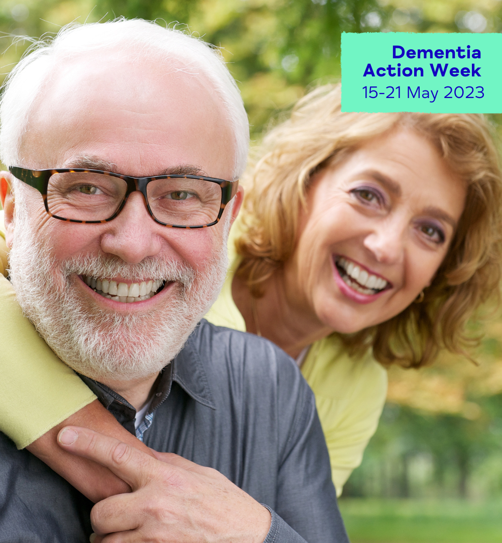 A man aged in his sixties with white hair and dark rimmed glasses, smiles towards the camera, whilst being hugged by his female companion who is also smiling. They are outdoors surrounded by greenery.