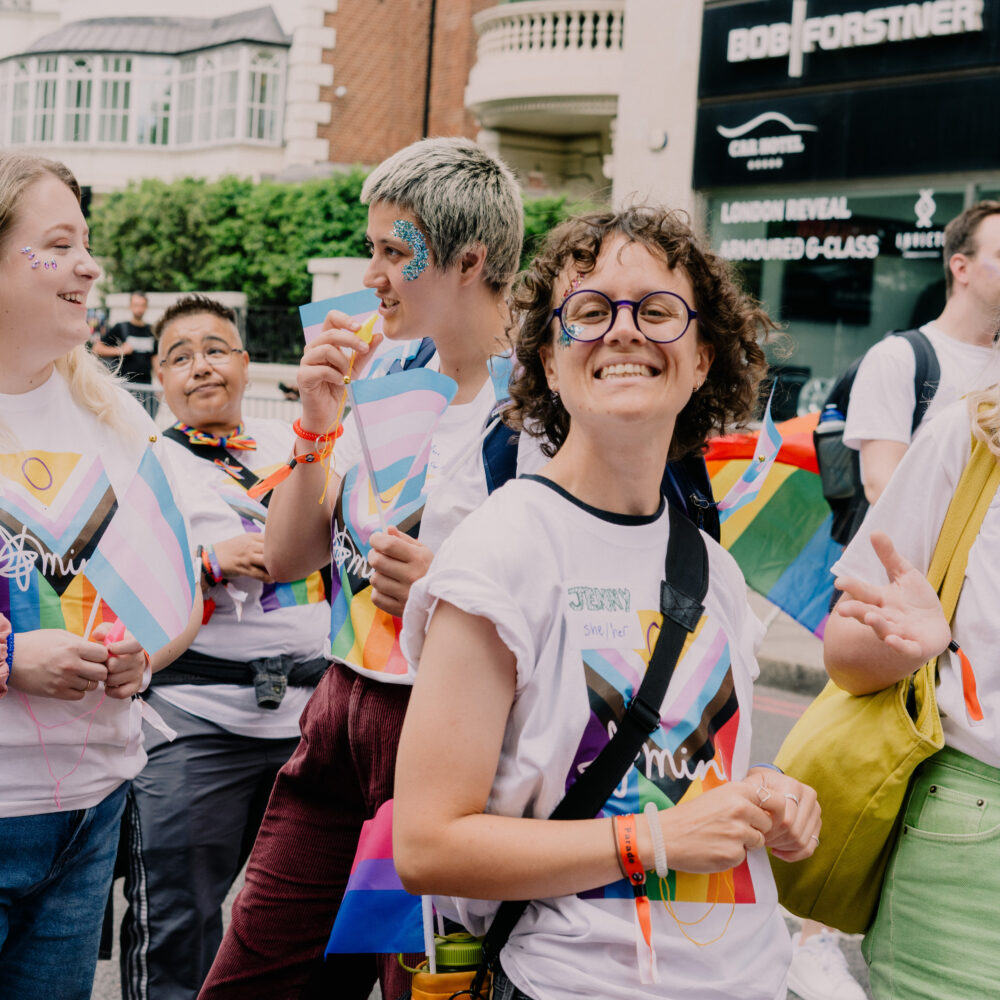 People wearing pride rainbow tshirts are particpating in a parade, smiling towards the camera. They have rainbow glitter on their faces.